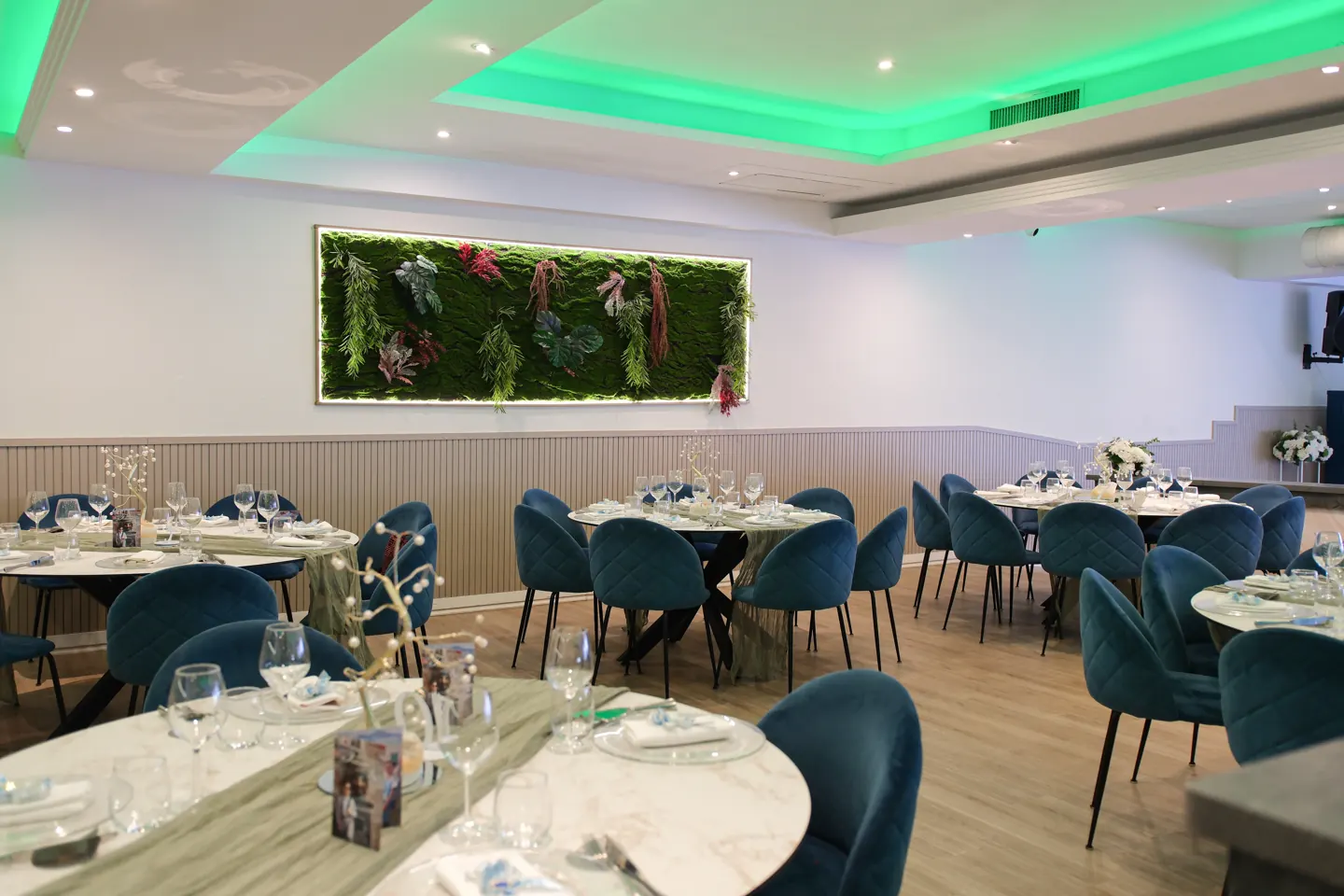 Sala apparecchiata con sedute blu, tavoli tondi, parete vegetale e luci verdi.
